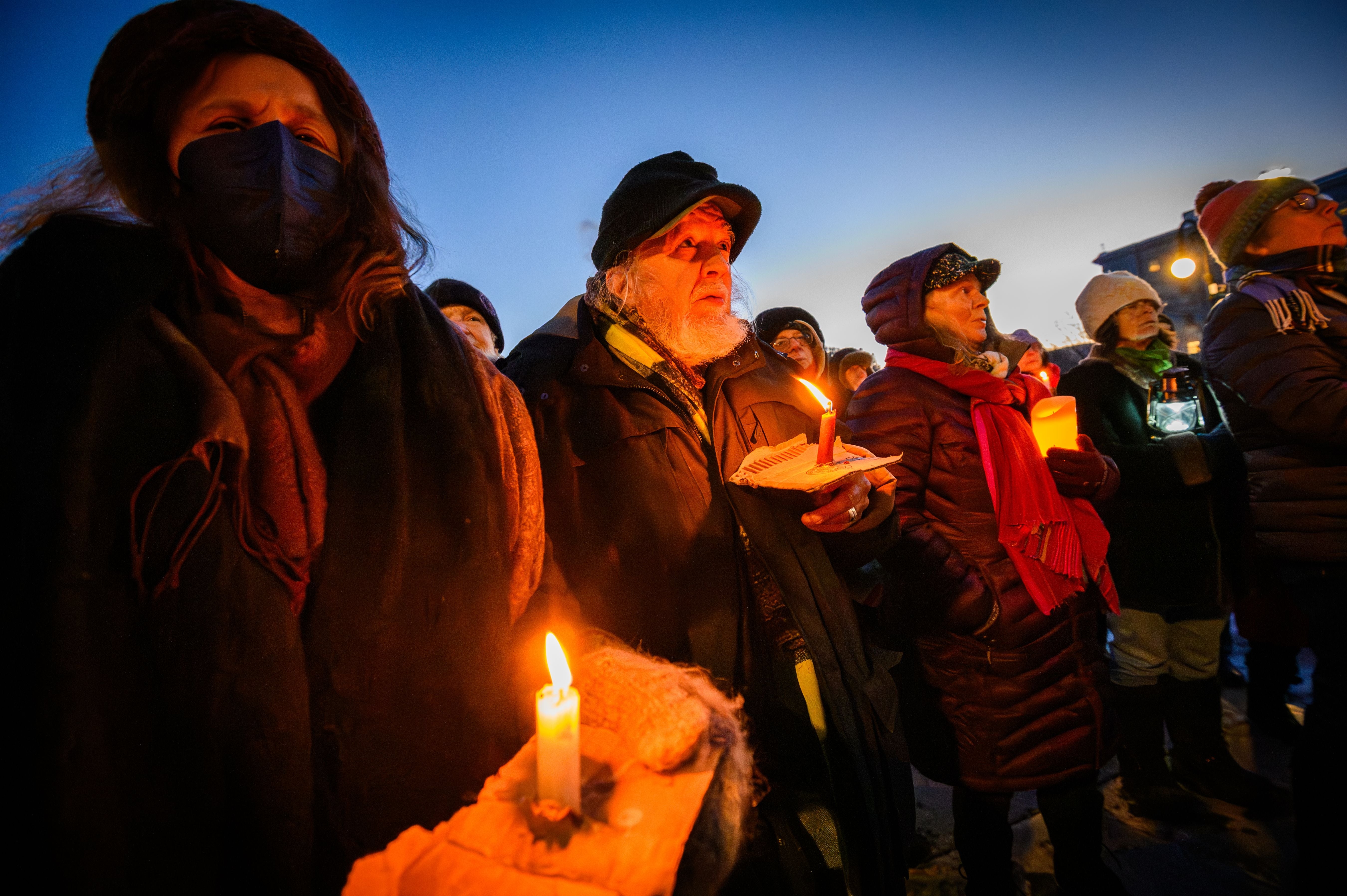 People lasting outdoors astatine dusk, holding lit candles during a vigil. They are dressed warmly with hats and scarves
