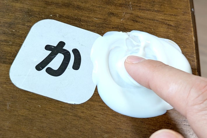 指がクリームを押している木目調の表面には「か」の文字があります。