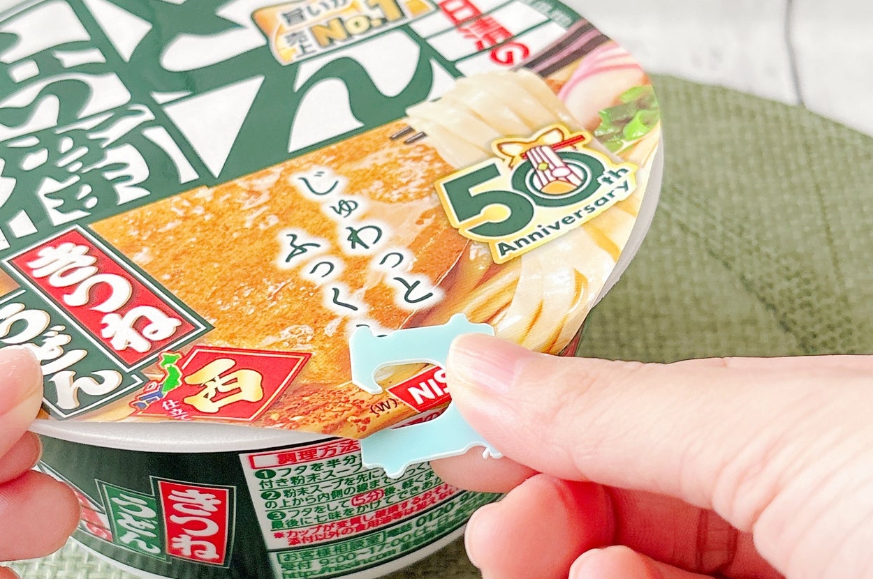 カップ麺の蓋に50周年ステッカー。手がシールを剥がそうとしている。蓋には商品名や「No.1」などのテキスト。