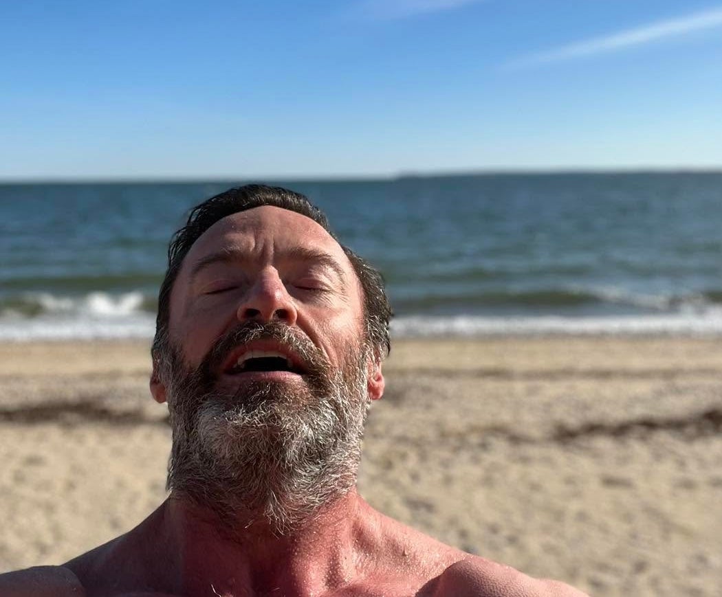 Hugh Jackman with his eyes closed sighing with relief on a beach with the waves in the background