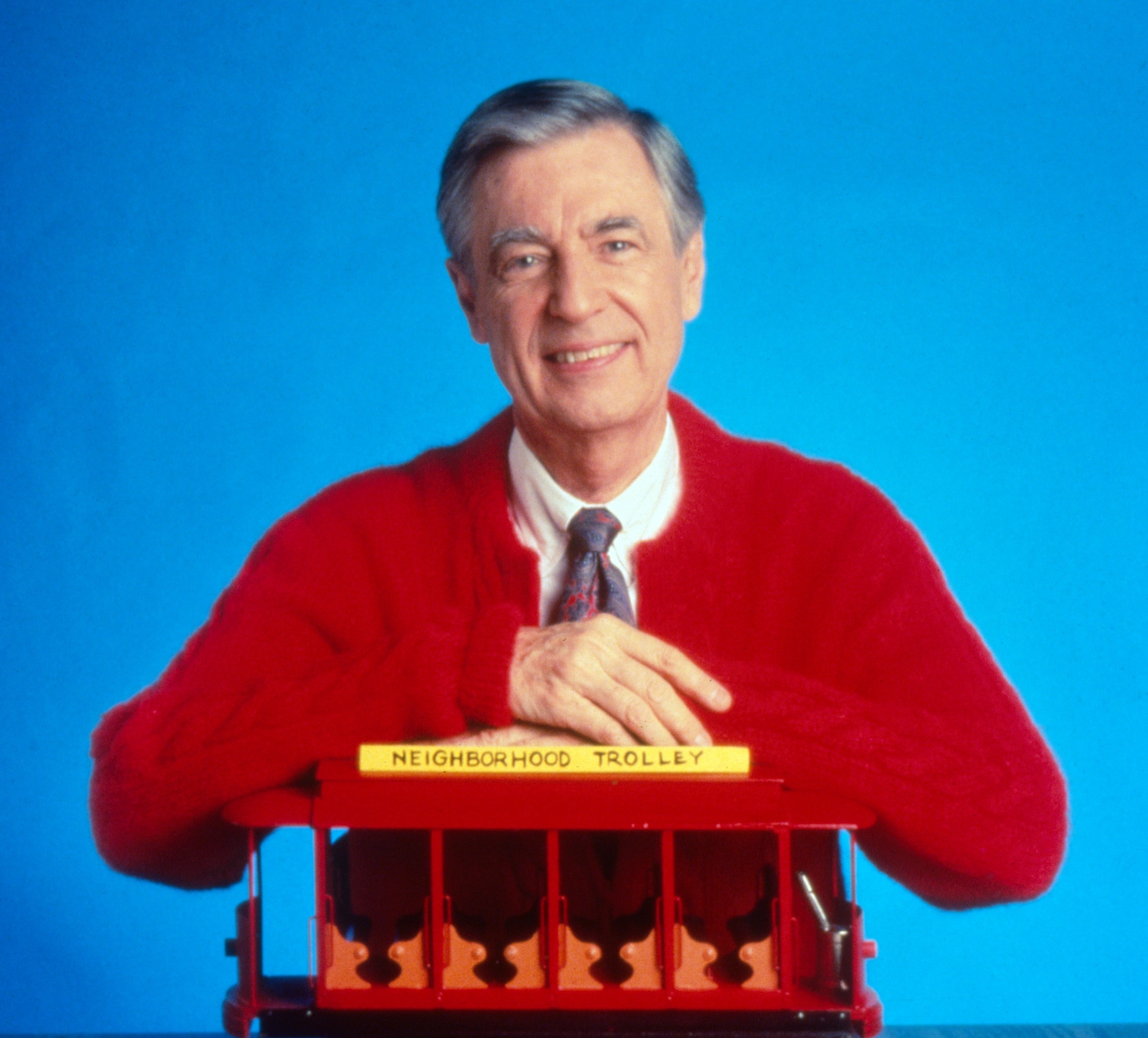 Mr. Rogers in his iconic red cardigan smiles as he leans on a model trolley