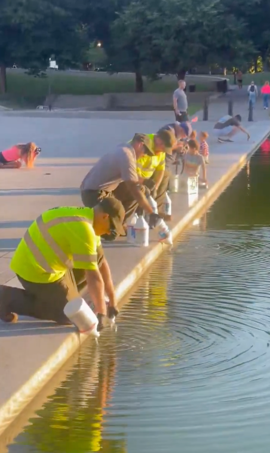 People successful high-visibility vests cleanable the borderline of a shallow nationalist h2o feature, utilizing buckets and gloves, nether a agleam sky