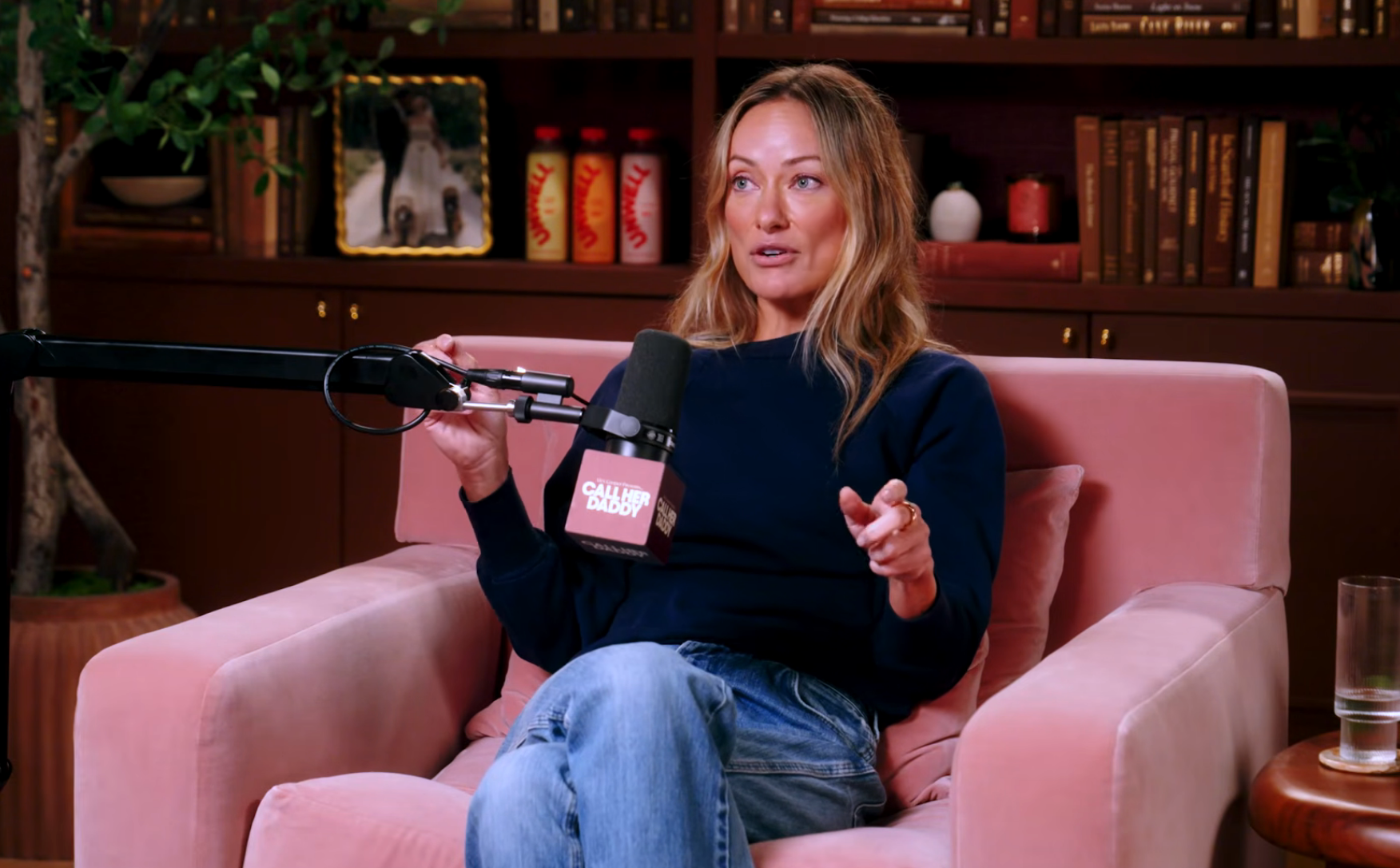 Person sitting successful  a podcast studio, speaking into a microphone, wearing a casual sweater and jeans, surrounded by books and decor