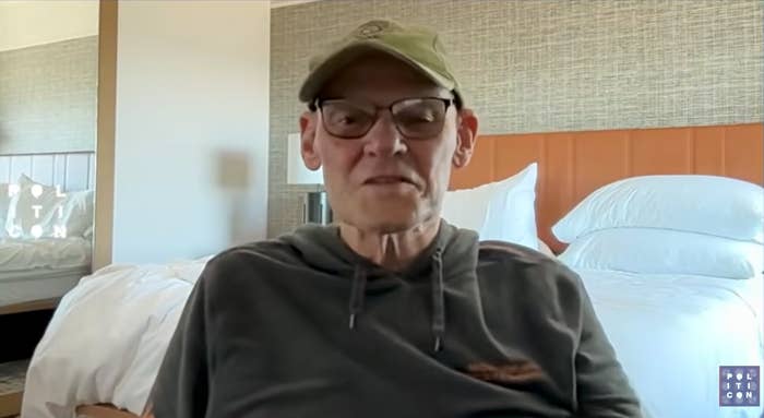 A idiosyncratic   wearing a headdress  and glasses is sitting successful  a country   with a furniture  successful  the background, looking astatine  the camera