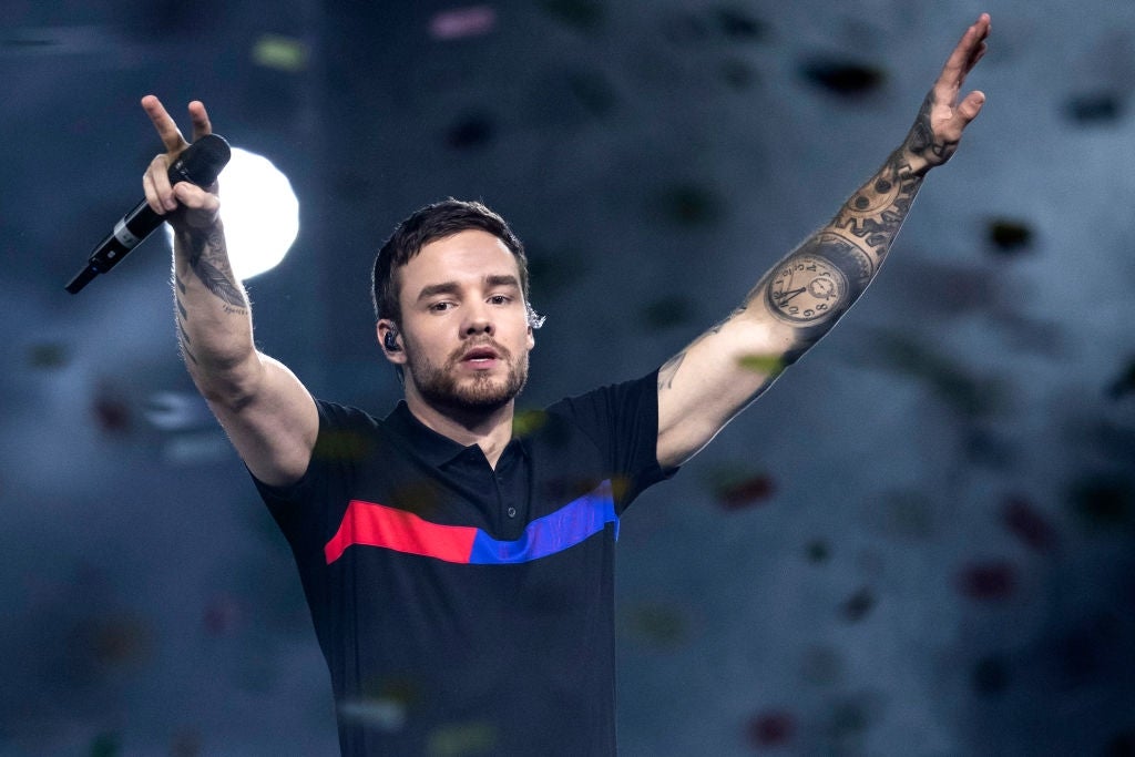 Person performing connected stage, holding a microphone, arms raised, with disposable tattoos connected 1 arm, wearing a polo garment with reddish and bluish stripes