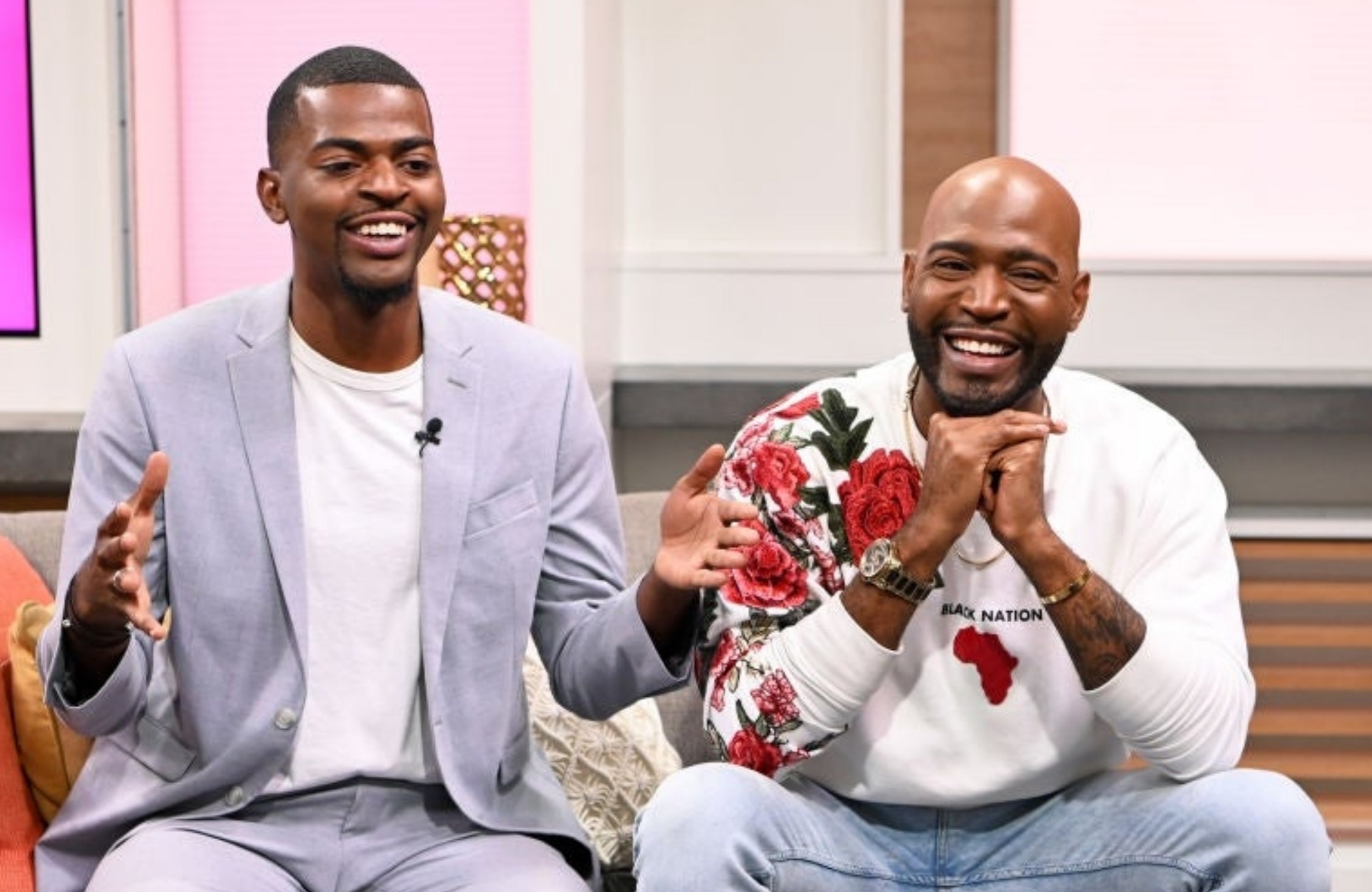 Two men sitting connected a couch, smiling and laughing during a speech show. One wears a suit, the different a casual sweatshirt with a floral design