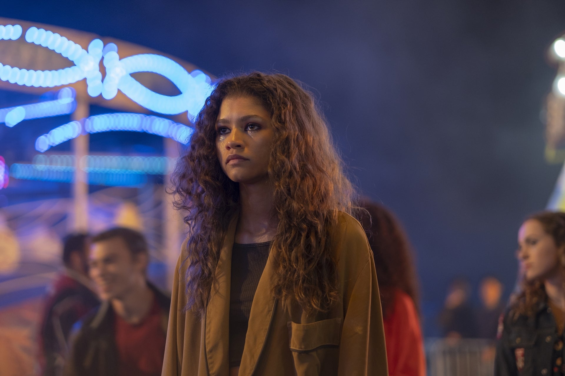 Person with curly hair in a casual jacket stands at a fairground, surrounded by vibrant lights and blurred people in the background