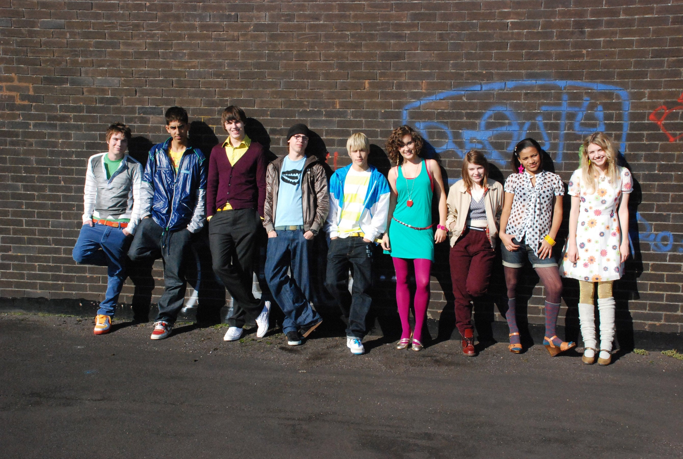 A group of nine young adults leaning against a brick wall, each displaying casual, eclectic fashion styles