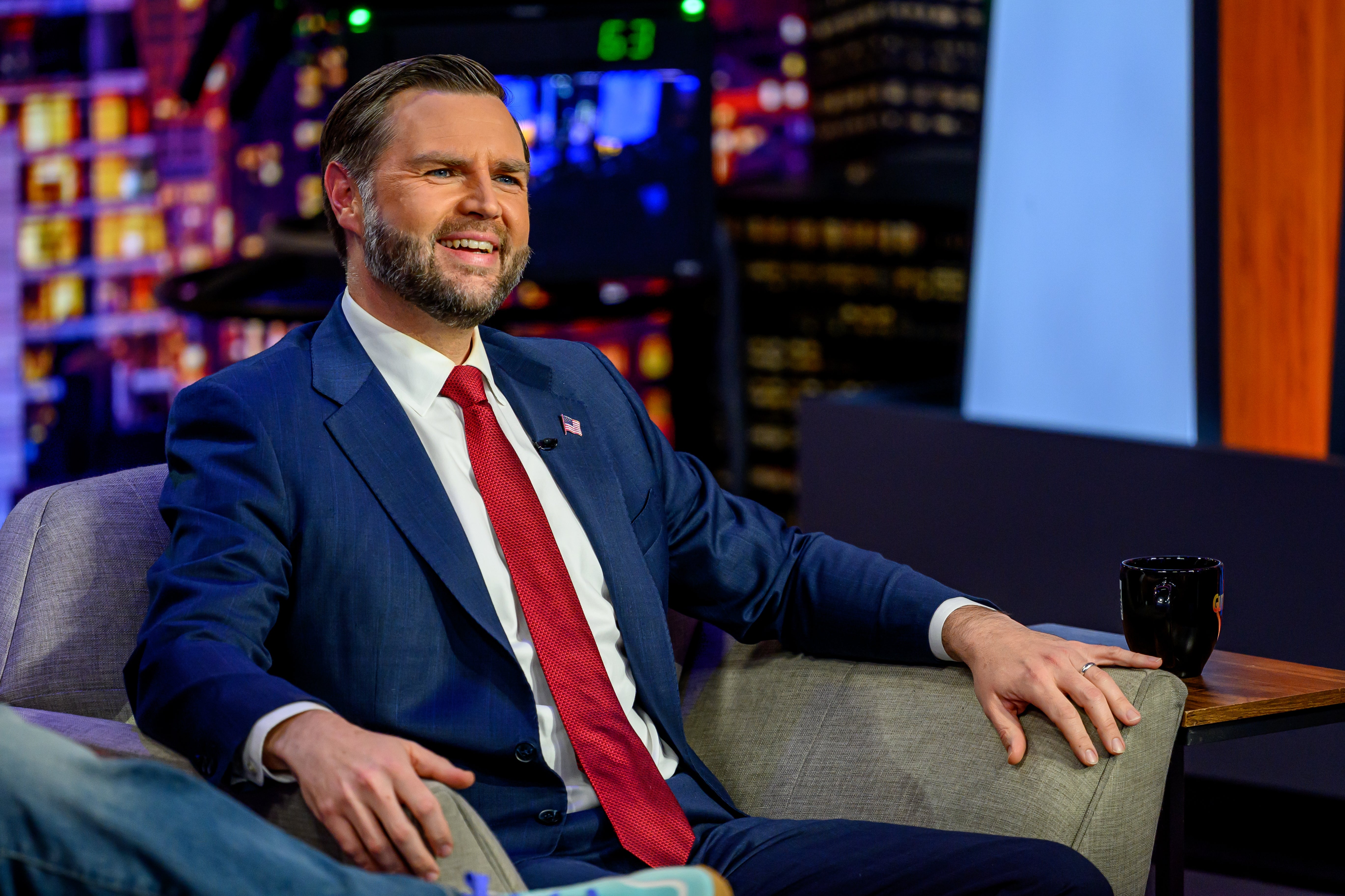 A idiosyncratic successful a suit and tie, sitting and smiling connected a speech amusement set, with a nighttime metropolis backdrop and a java mug beside them
