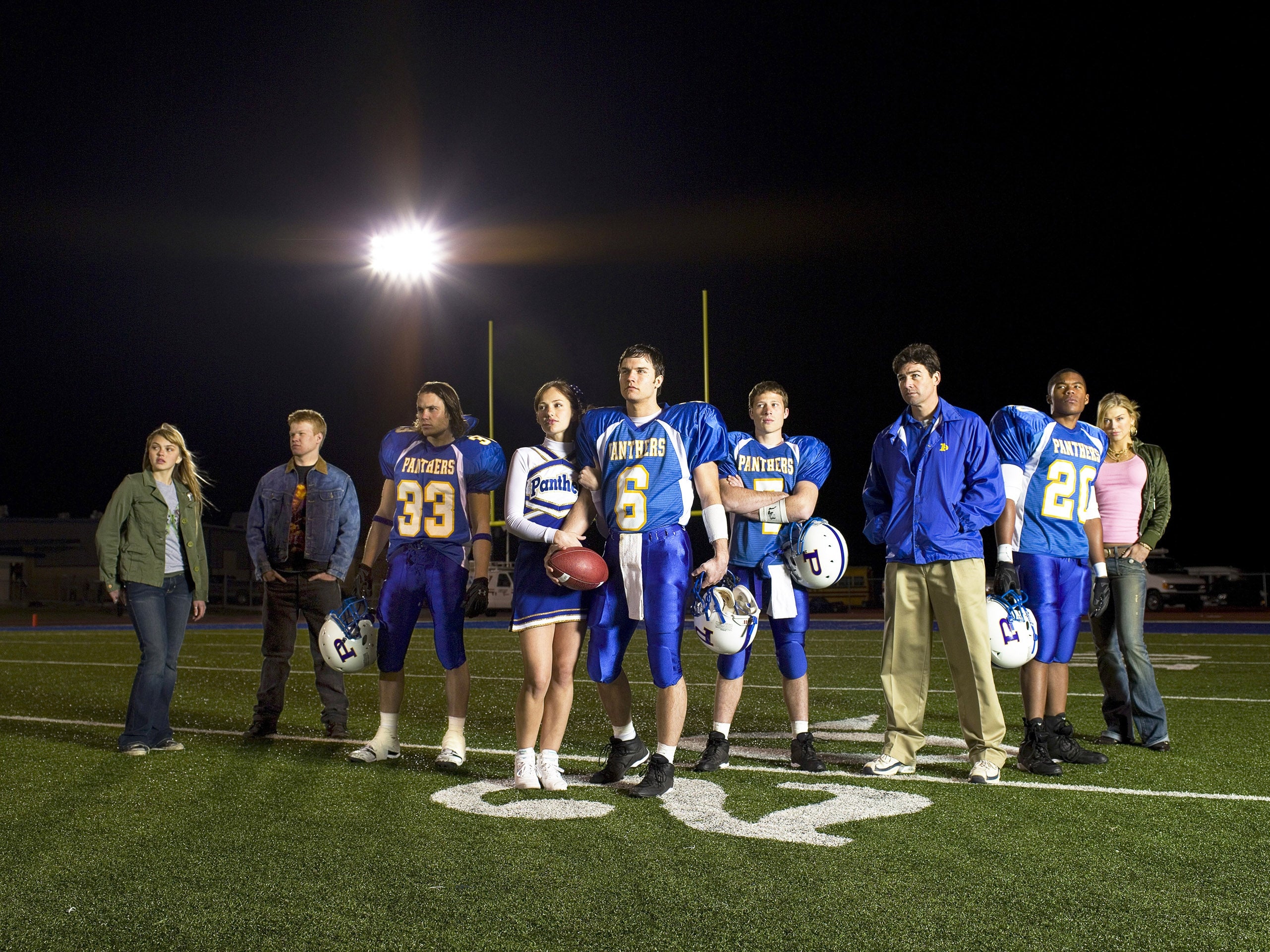 Cast of a high school football-themed TV show stands on a field, some in jerseys, others in casual attire, under stadium lights