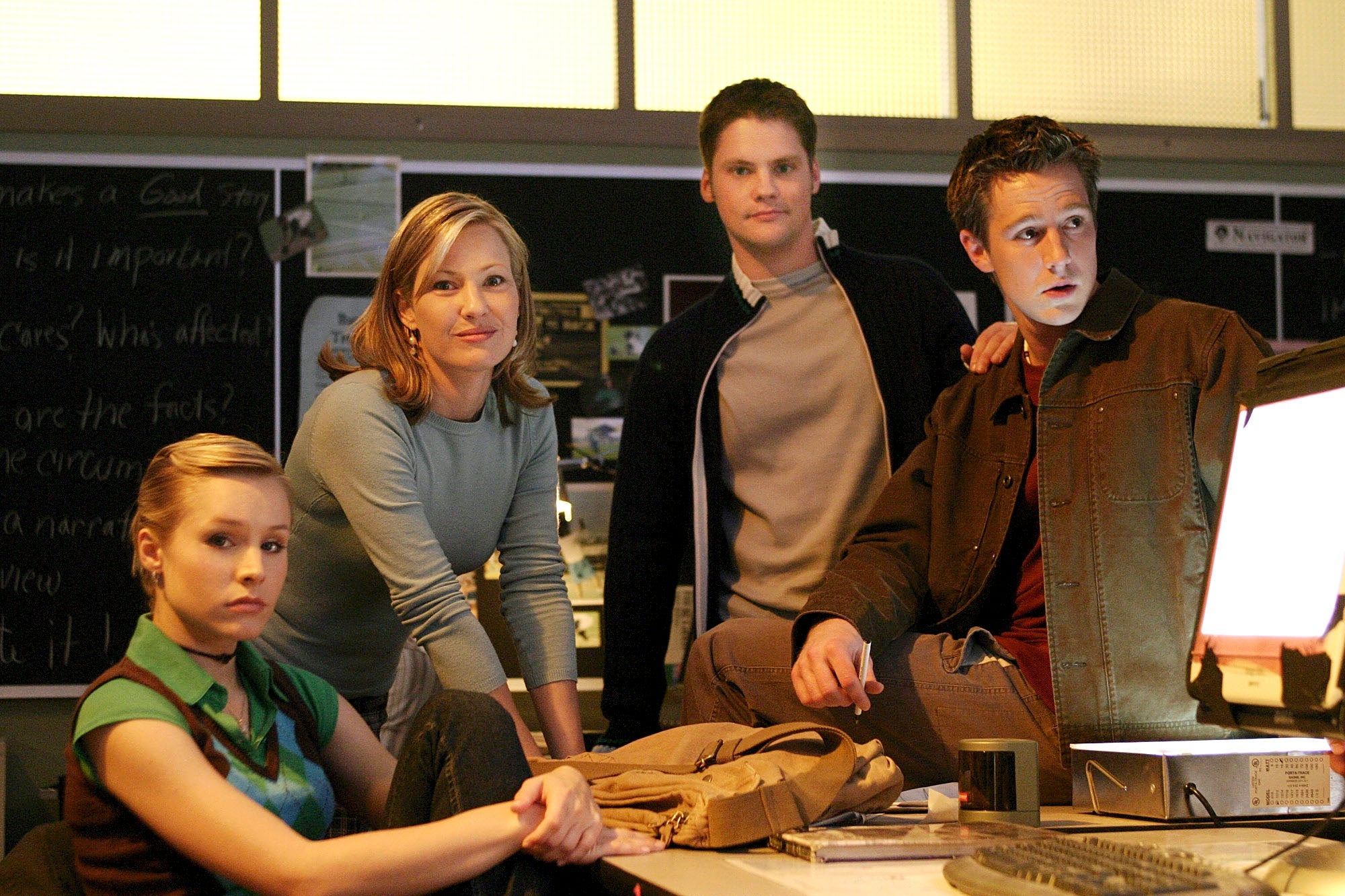 Four people are gathered around a desk in a detective office setting, looking intently. They appear focused and engrossed in their environment