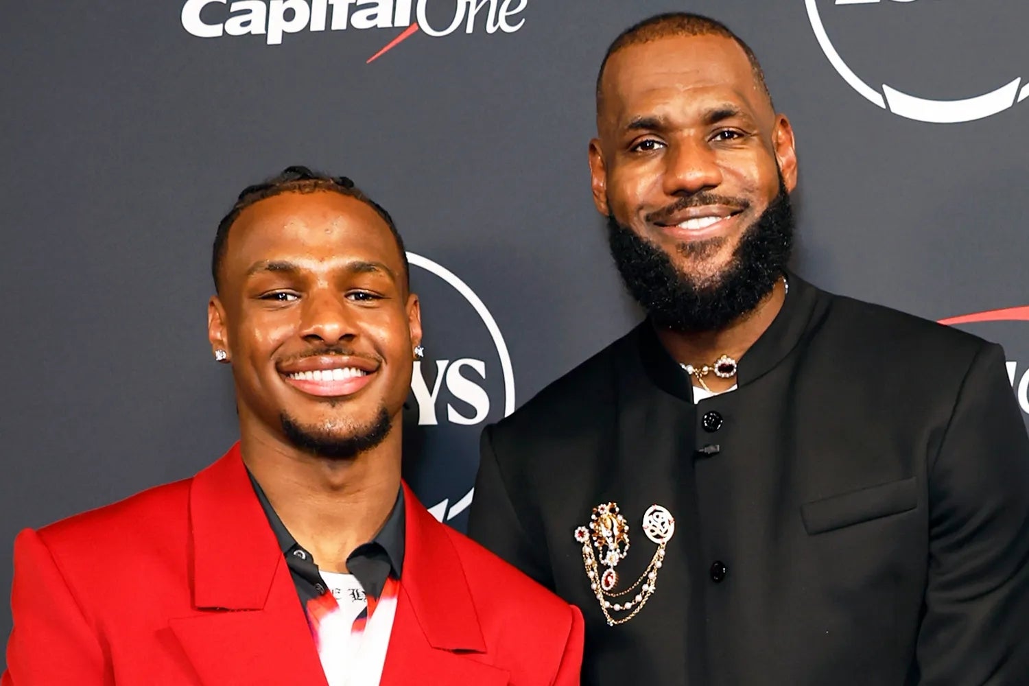 Two individuals astatine a ceremonial event; 1 successful a reddish overgarment and the different successful a achromatic suit with decorative pins. They are smiling astatine the camera