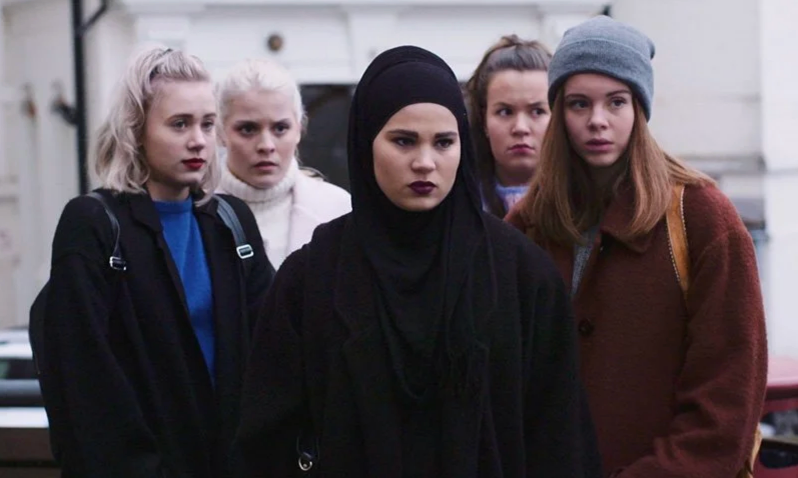 A group of five individuals stands together, each displaying a distinct fashion style, including casual and modest attire, possibly part of a scene in TV or movies