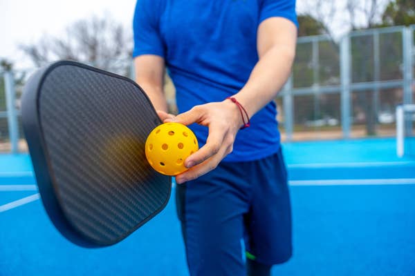 Person holding a pickleball paddle and yellow ball on an outdoor court