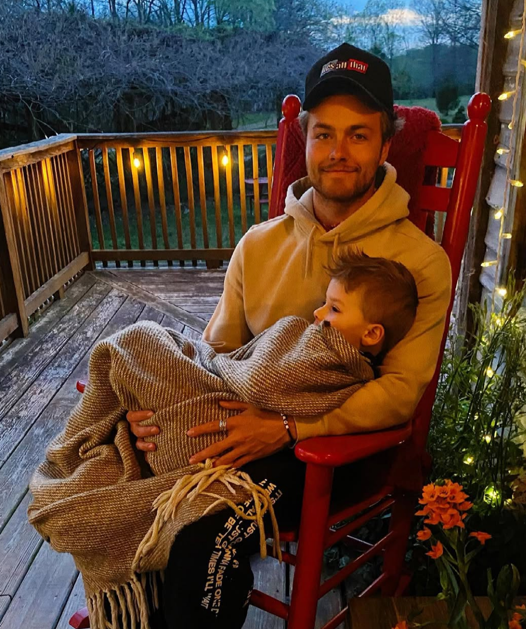 A idiosyncratic wearing a hoodie and headdress sits connected a structure successful a rocking chair, holding a kid wrapped successful a blanket. They look contented and relaxed
