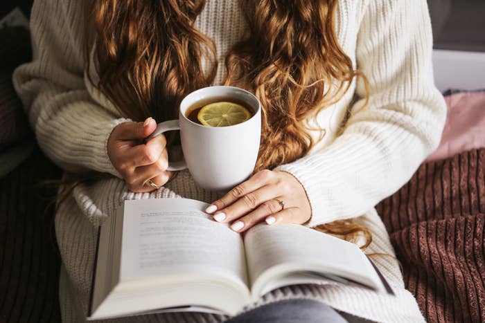 Person with agelong  hairsbreadth  reads a book, holding a mug with a citrus  slice. Sweater and cozy mounting  suggest   a relaxed speechmaking  moment