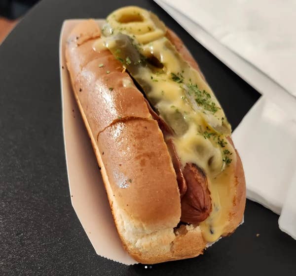 Hot dog topped with melted cheese, pickles, and herbs on a bun, served on a white tray