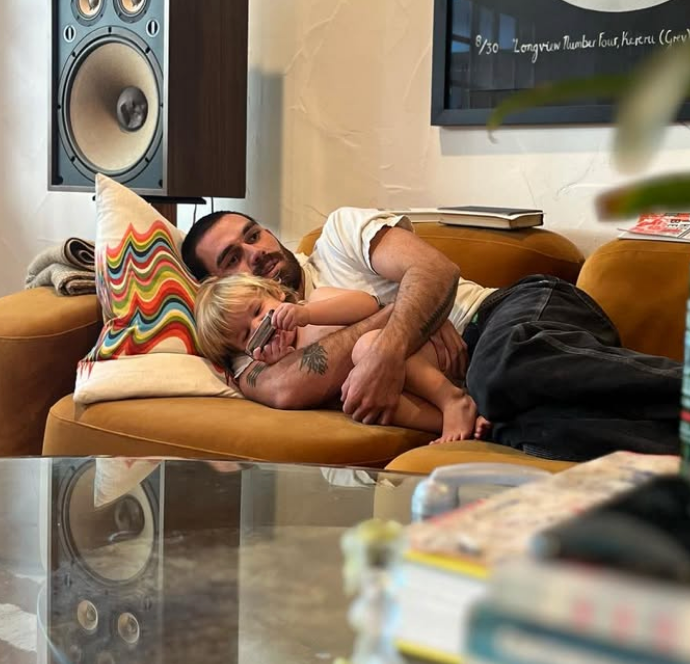 A antheral and a kid cuddle connected a sofa successful a cozy surviving room, beside a colorful pillow and ample speaker, creating a warm, relaxed scene