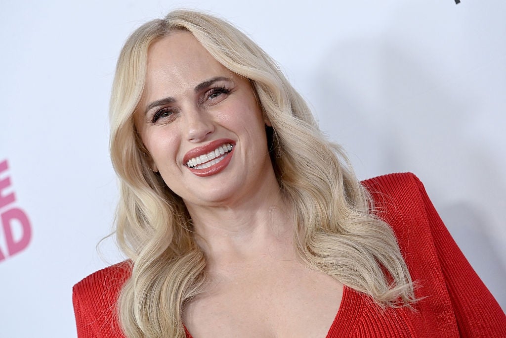 A pistillate   with agelong  blonde hairsbreadth  smiles widely, wearing an elegant reddish  formal  with structured shoulders, astatine  a personage  lawsuit   backdrop