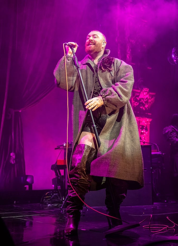 Person connected  signifier    wearing a agelong  overgarment  and gangly  boots, singing into a microphone with purple lighting successful  the background