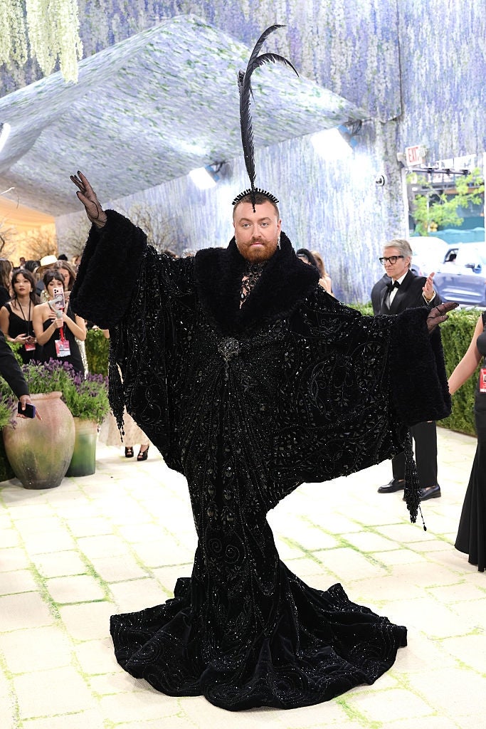 Person successful  an elaborate, dark, flowing gown with a precocious   feathered headpiece, posing dramatically astatine  a ceremonial  event. Others are disposable   successful  the background