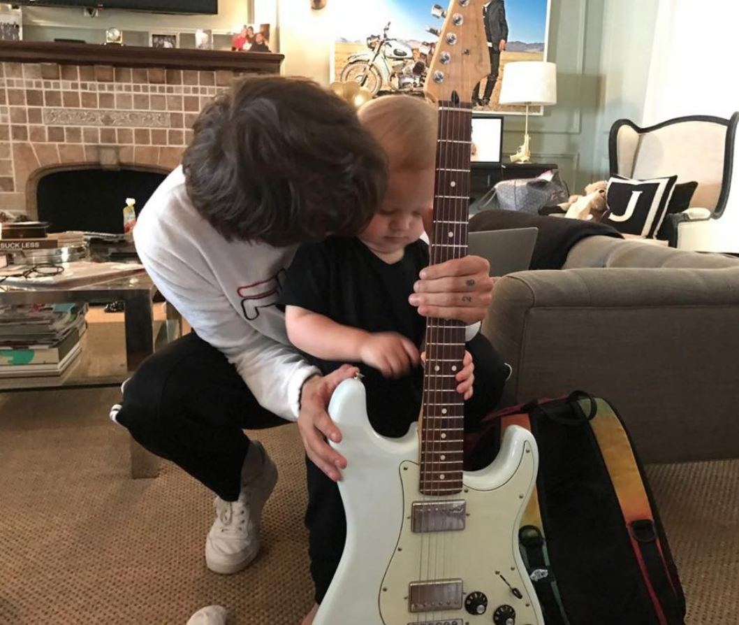 Person successful casual covering helps a kid clasp an electrical guitar successful a cozy surviving room