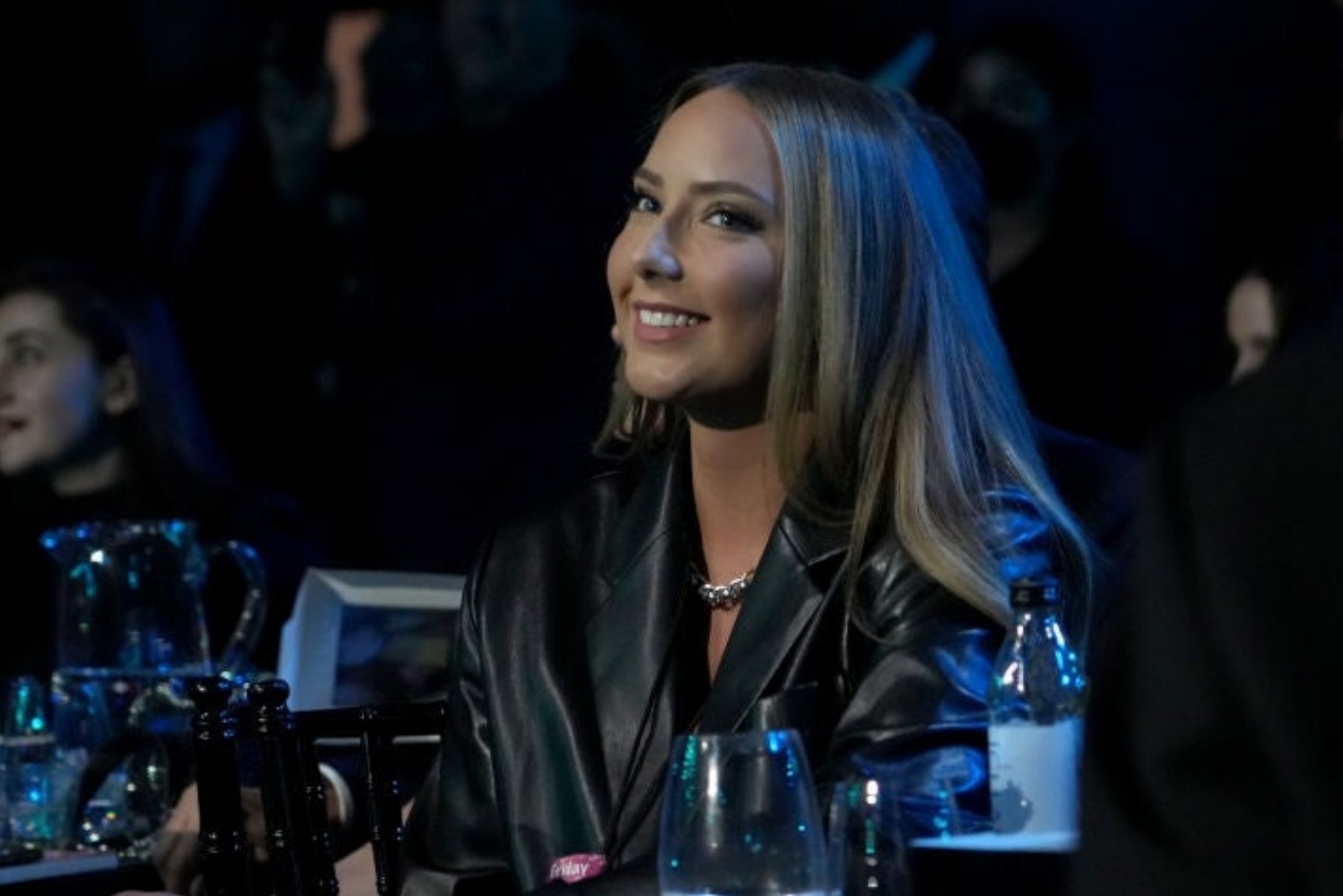Woman sitting astatine an lawsuit successful a chic leather jacket, smiling; guests are disposable successful the dimly lit background
