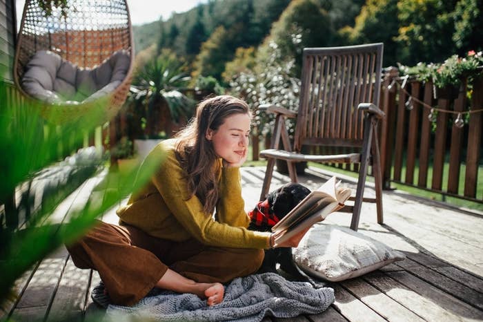 A pistillate   sits connected  a woody  platform  speechmaking  a publication  with a canine  beside her, surrounded by quality  and outdoor furniture