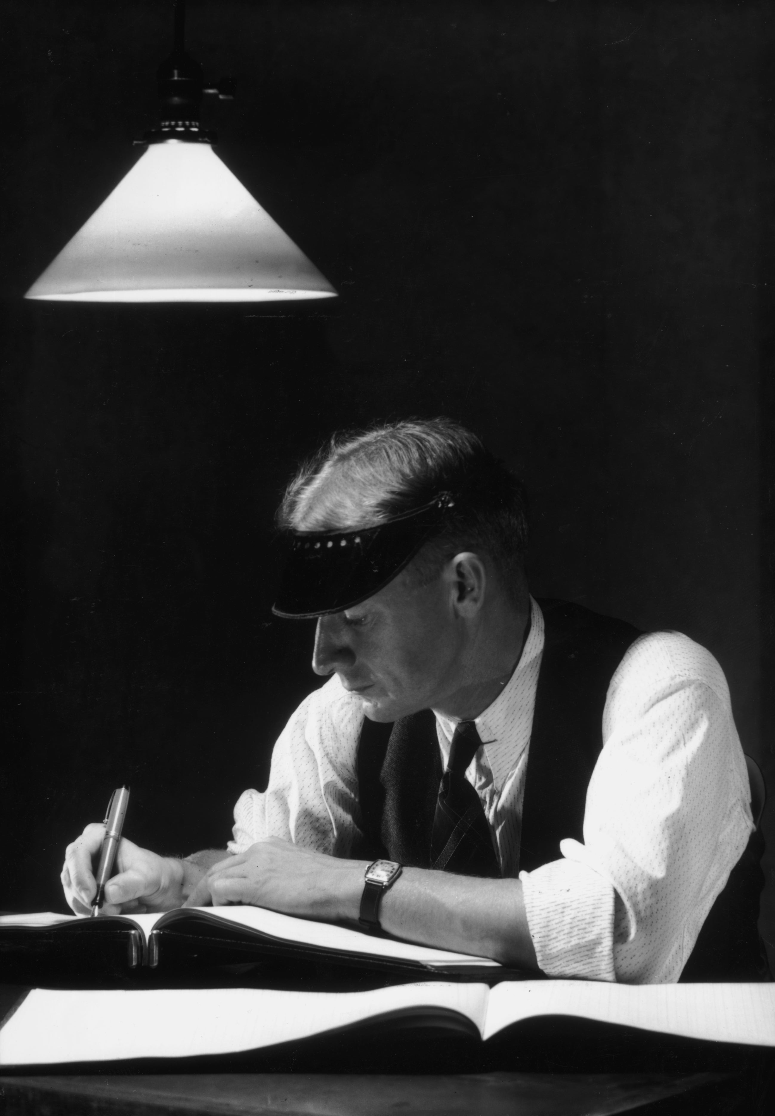 A antheral wearing a visor writes astatine a table nether a hanging lamp, surrounded by unfastened books, creating a focused and studious atmosphere