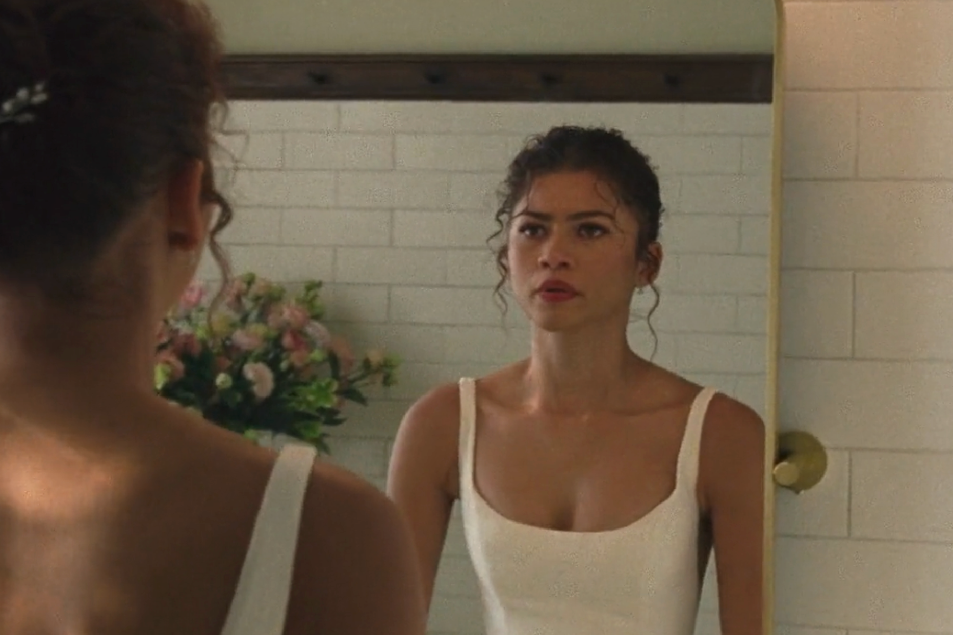 A woman in a sleeveless wedding dress looks at herself in a mirror, with flowers in the background