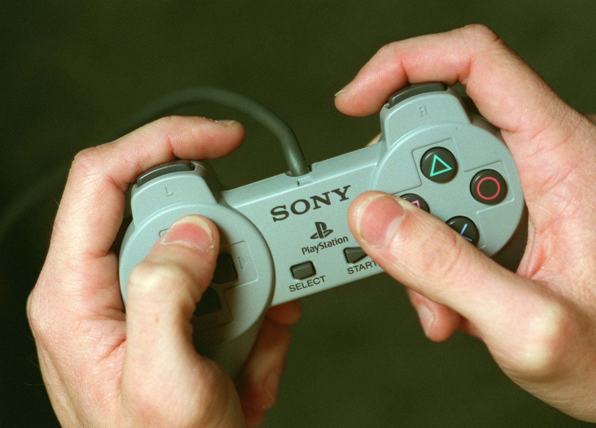 Hands holding a classical Sony PlayStation controller, pressing buttons with thumbs, suggesting retro gaming nostalgia