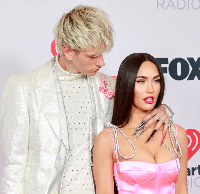 Two individuals on a red carpet. One wears a white suit with stud details and the other wears a pink dress