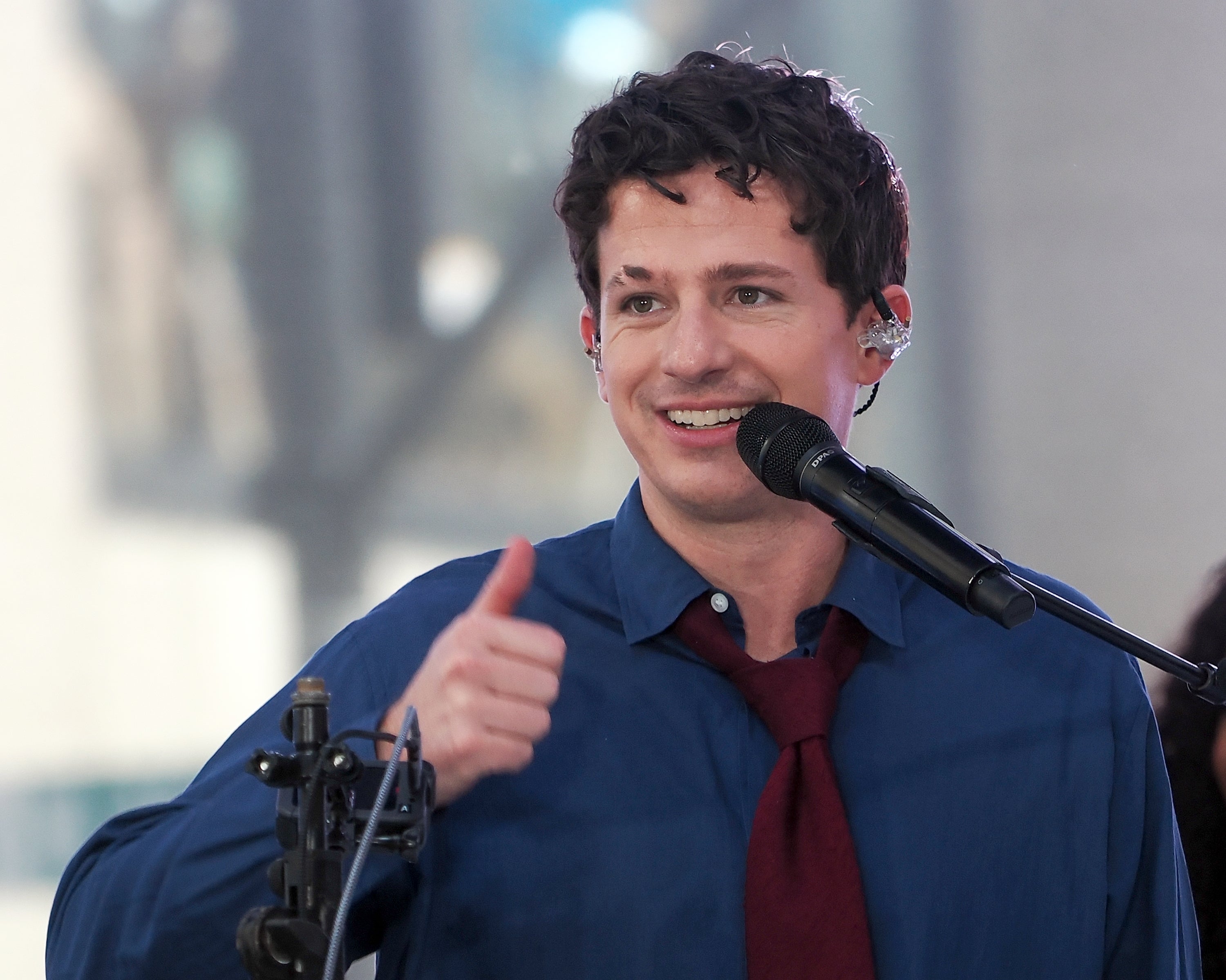 Person in front of a microphone wearing a button-up shirt and tie smiles and flashes a thumbs up