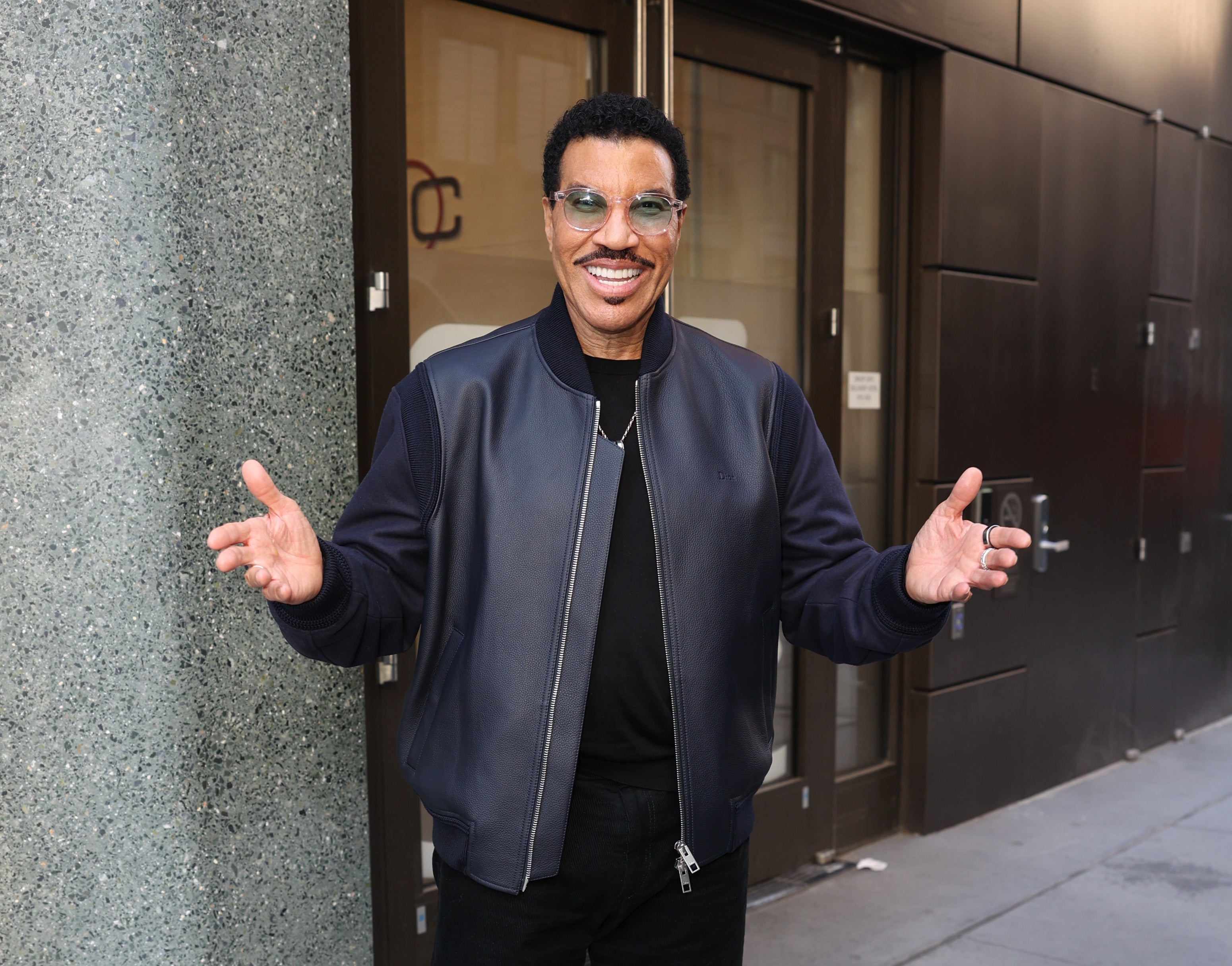 Man wearing a casual jacket and sunglasses smiling with open arms outside a building