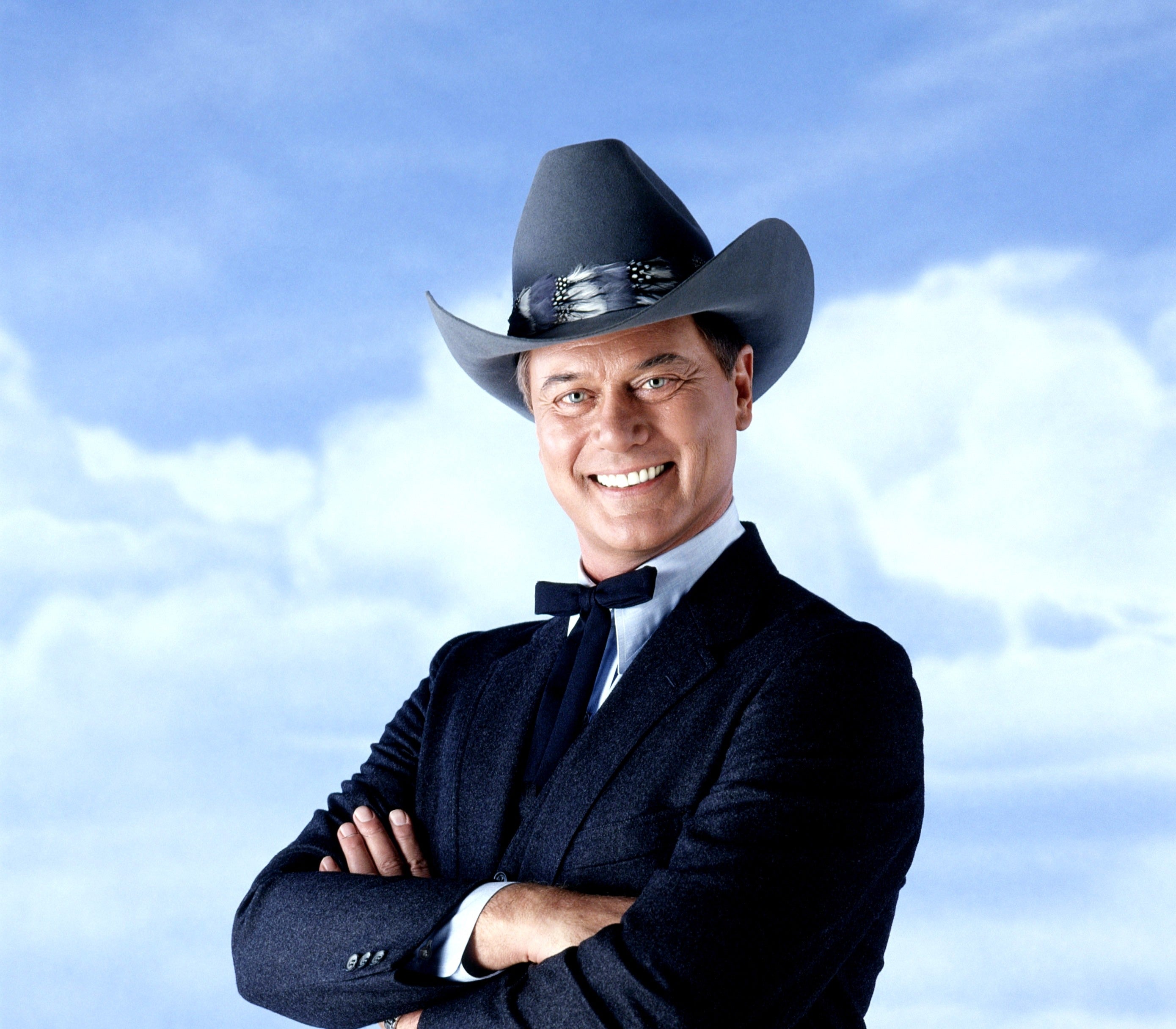 Larry Hagman in a suit and a large cowboy hat smiles with arms crossed against a sky background for a Dallas photoshoot