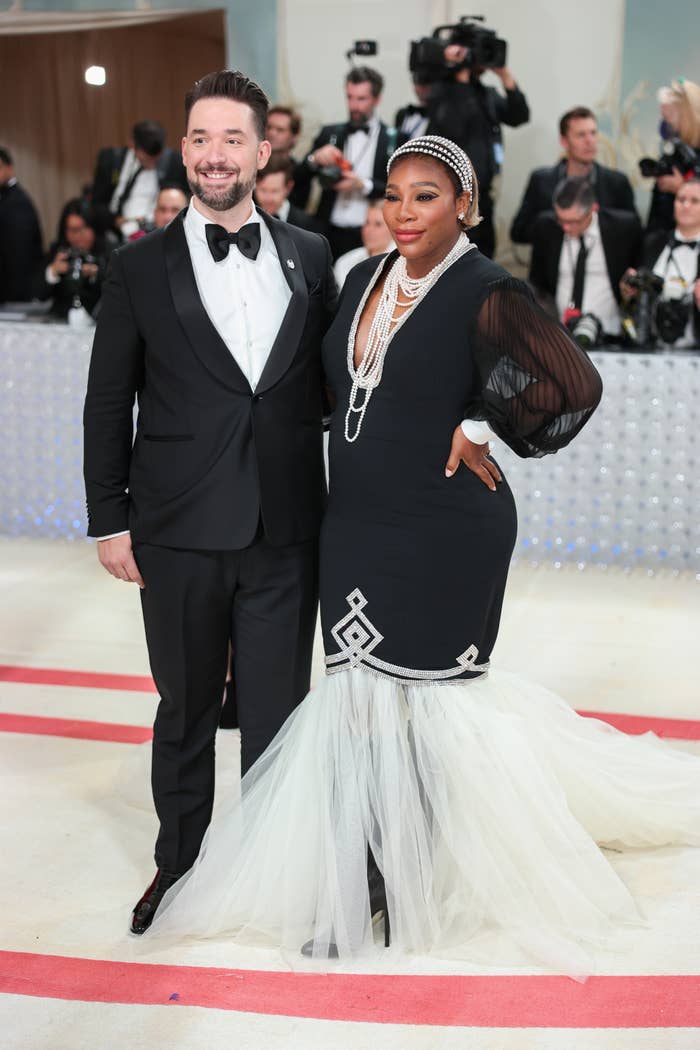 Alexis Ohanian wears a tuxedo and Serena Williams wears a fitted gown with sheer sleeves, pearl jewelry, and a tulle train