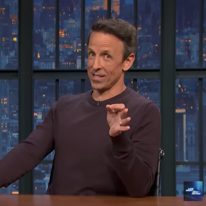 Person gesturing while sitting at a desk in a late-night talk show set
