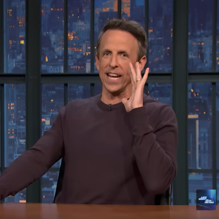 Person on nighttime TV set, gesturing with one hand near face, engaged in conversation