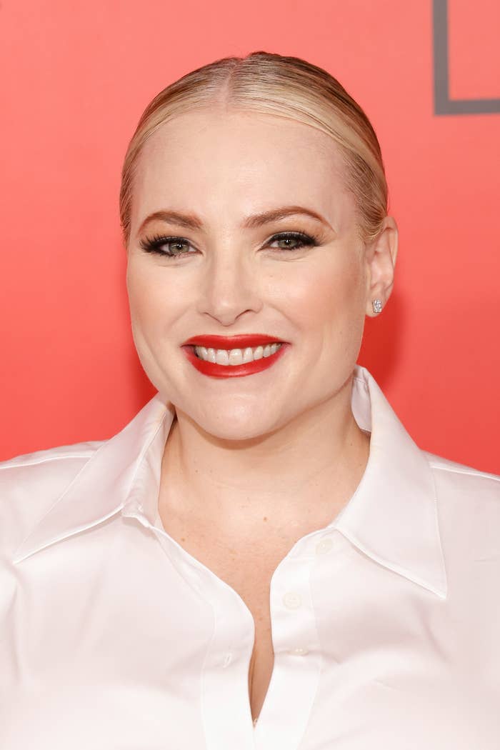 Person smiling in front of a red backdrop, wearing a white shirt with sleek, pulled-back hair and bold red lipstick