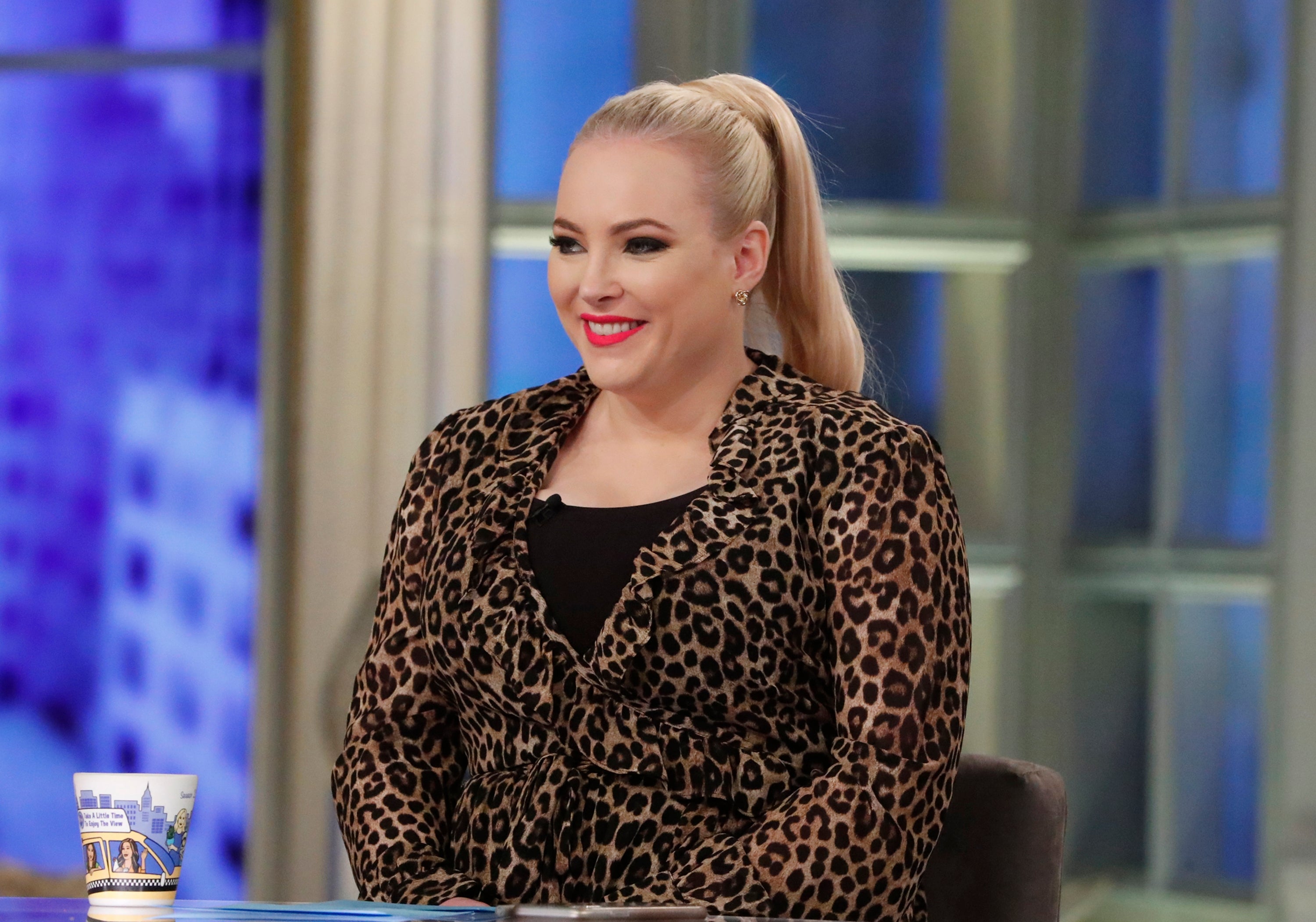 Person smiling on a talk show set, wearing a leopard-print outfit, seated at a table with a paper cup