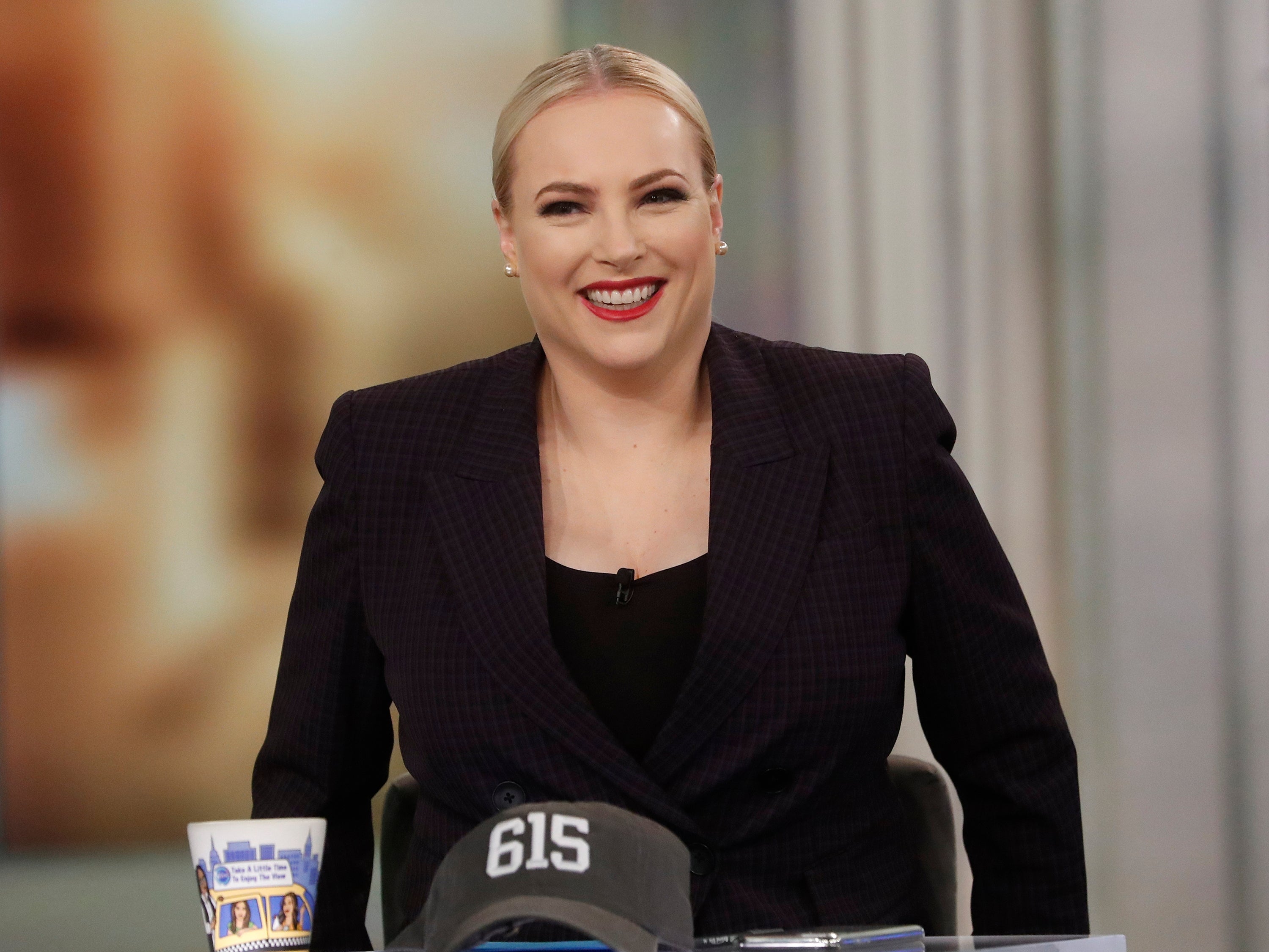 A smiling woman in a dark suit jacket sits at a desk with a mug and cap, appearing in a broadcast setting