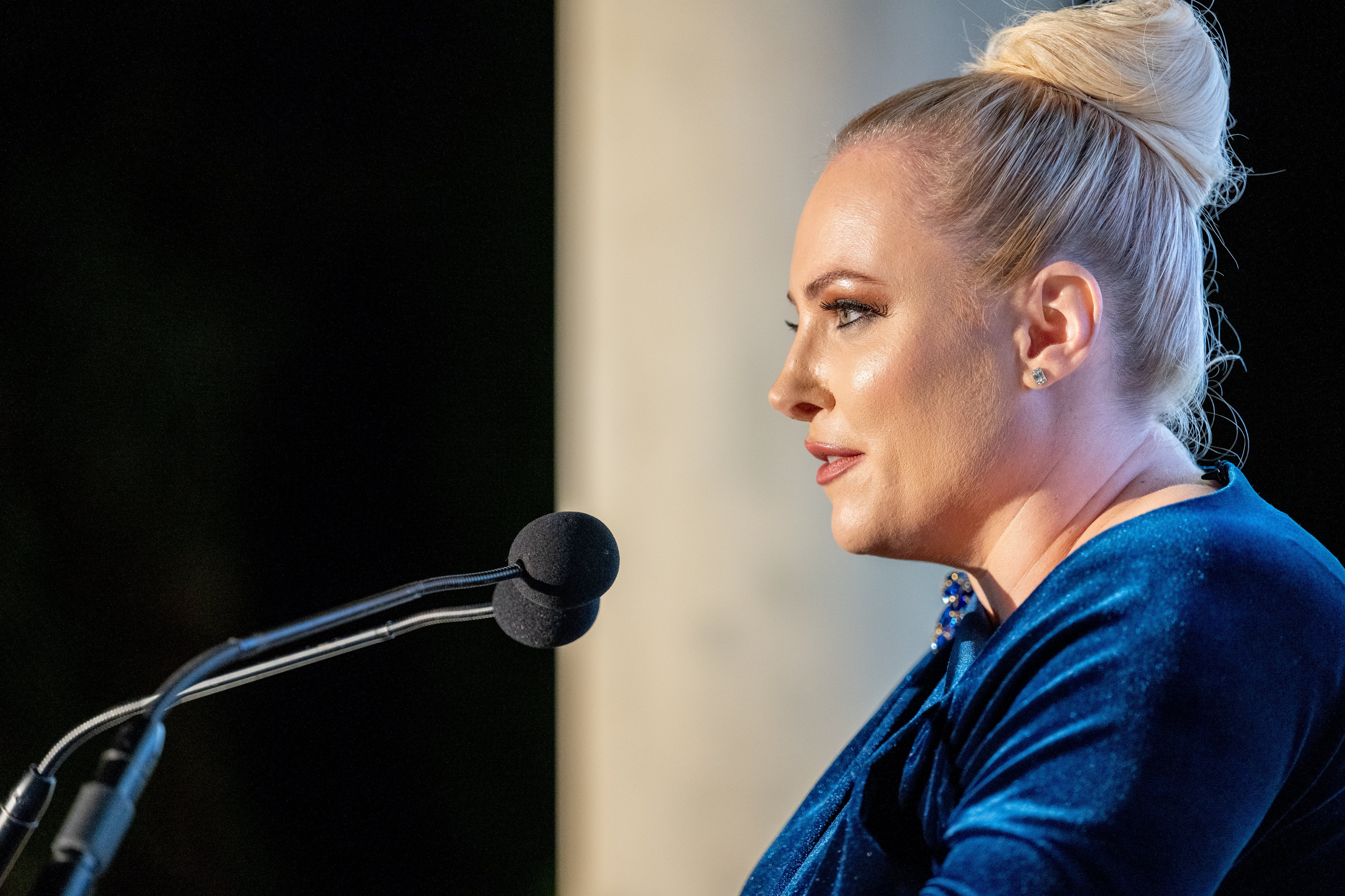 Person speaking at a podium, wearing an elegant velvet dress with hair in a bun, possibly at a formal event