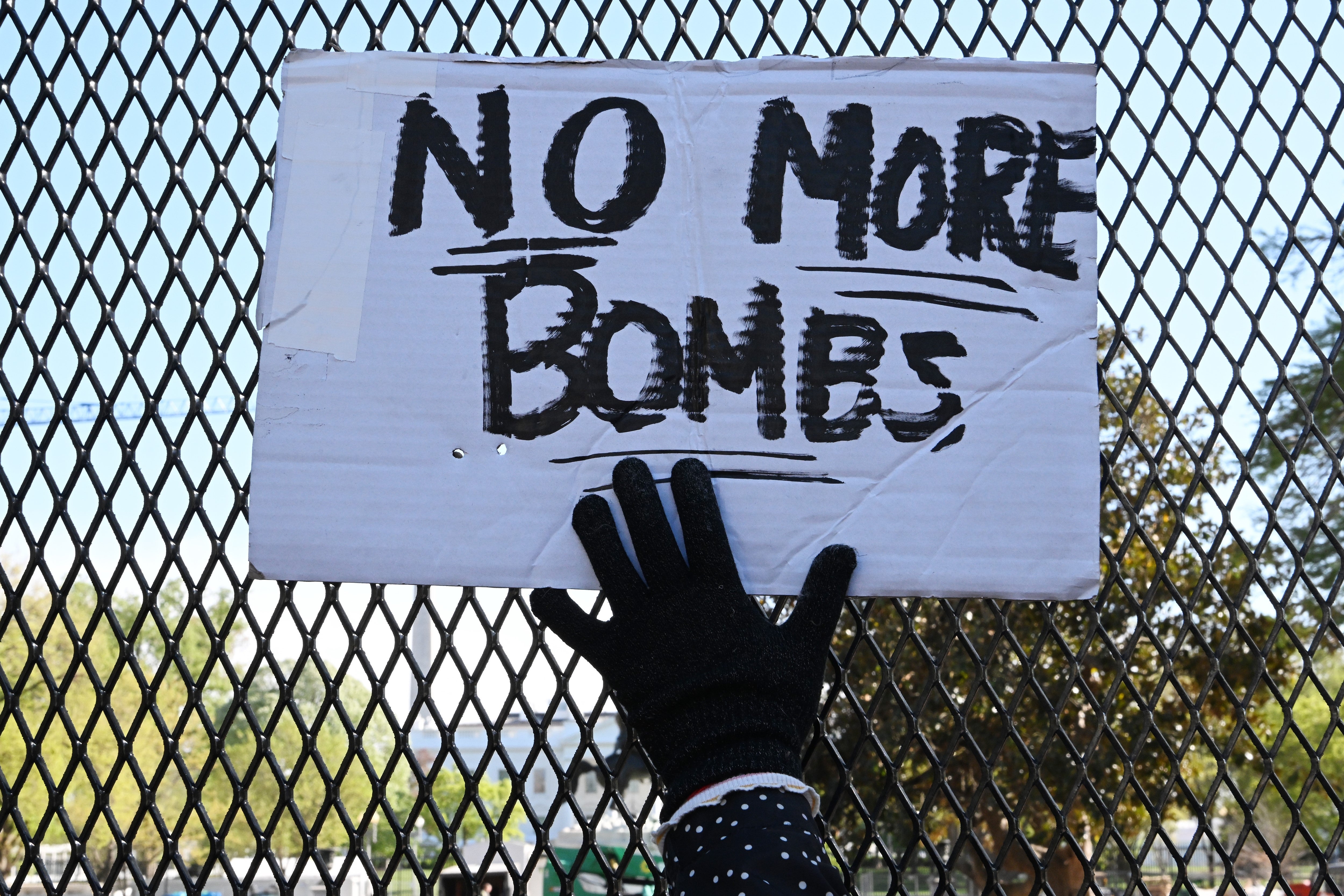 A gloved manus holds a motion speechmaking "No More Bombs" against a chain-link obstruction background