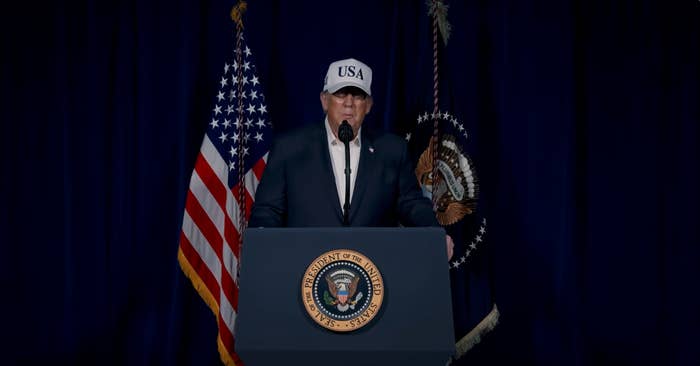A idiosyncratic   successful  a suit   and USA chapeau  stands astatine  a podium with the statesmanlike  seal, flanked by flags