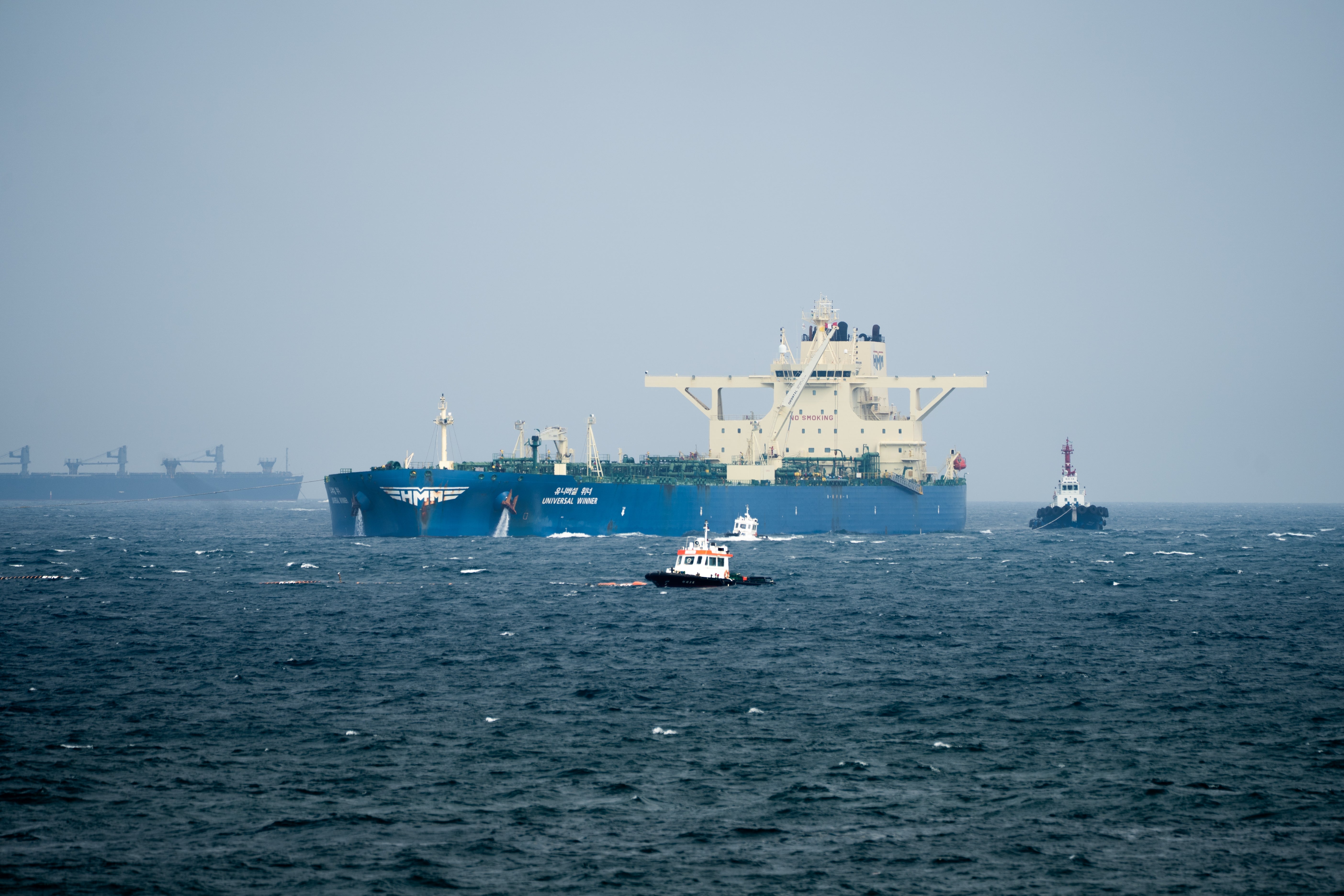 Large cargo vessel connected the water accompanied by smaller boats, nether a wide sky