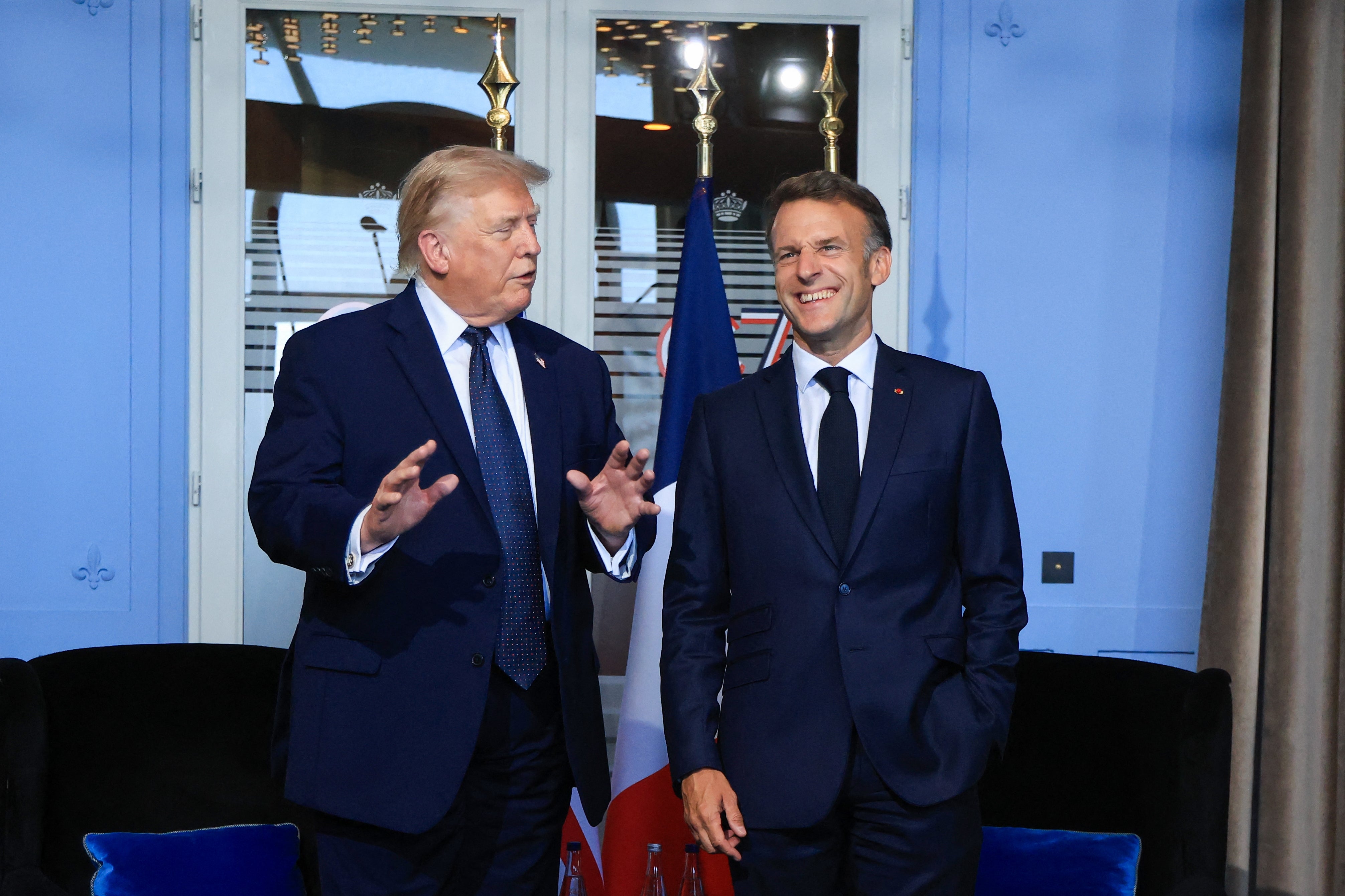 Two men successful suits basal together, 1 gesturing with hands. They look to beryllium successful speech successful a ceremonial mounting with flags successful the background