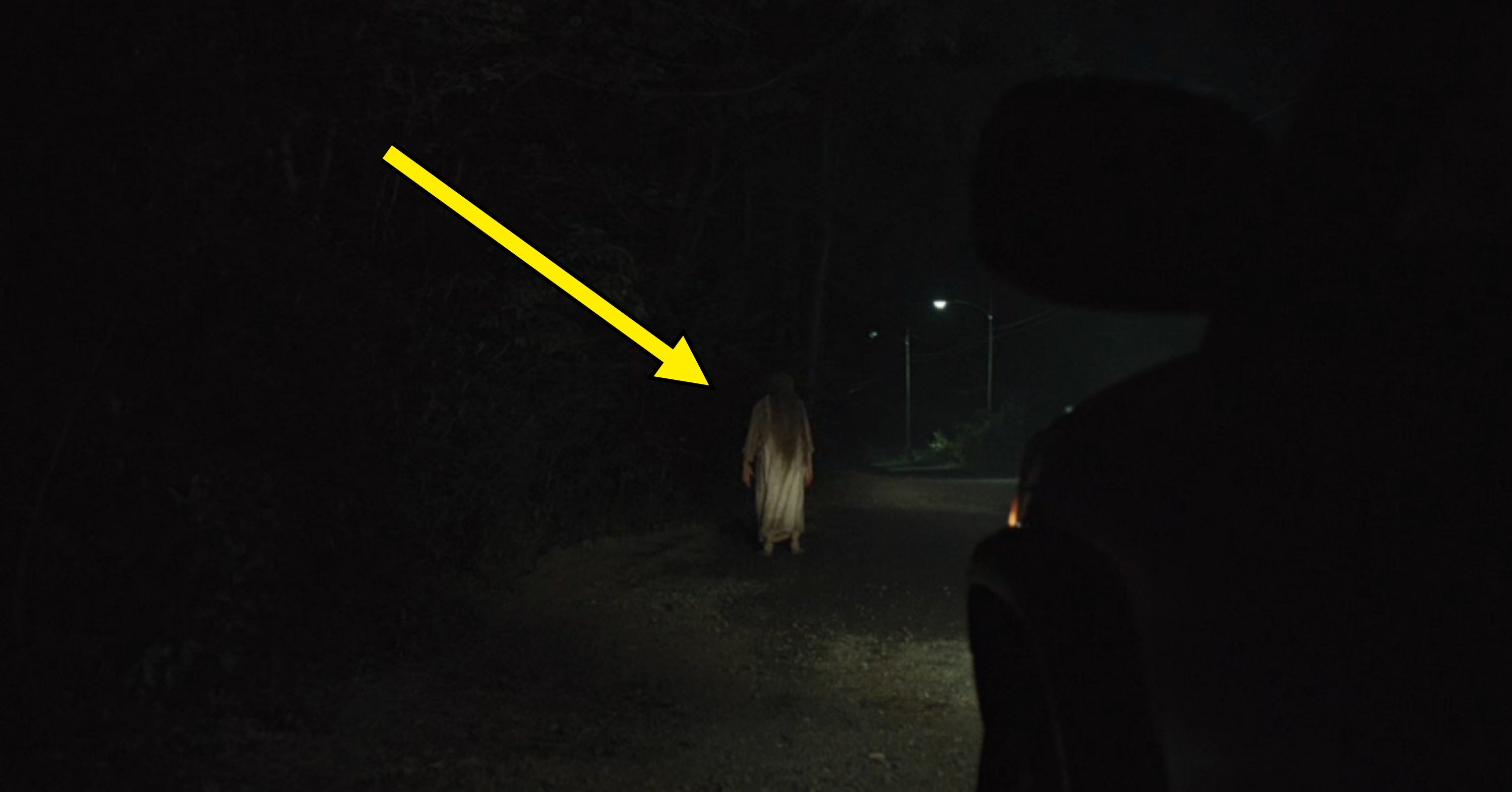 Person successful  agelong  cloak walking connected  dimly lit roadworthy  astatine  night, viewed from a car. Mysterious and atmospheric country   with minimal lighting