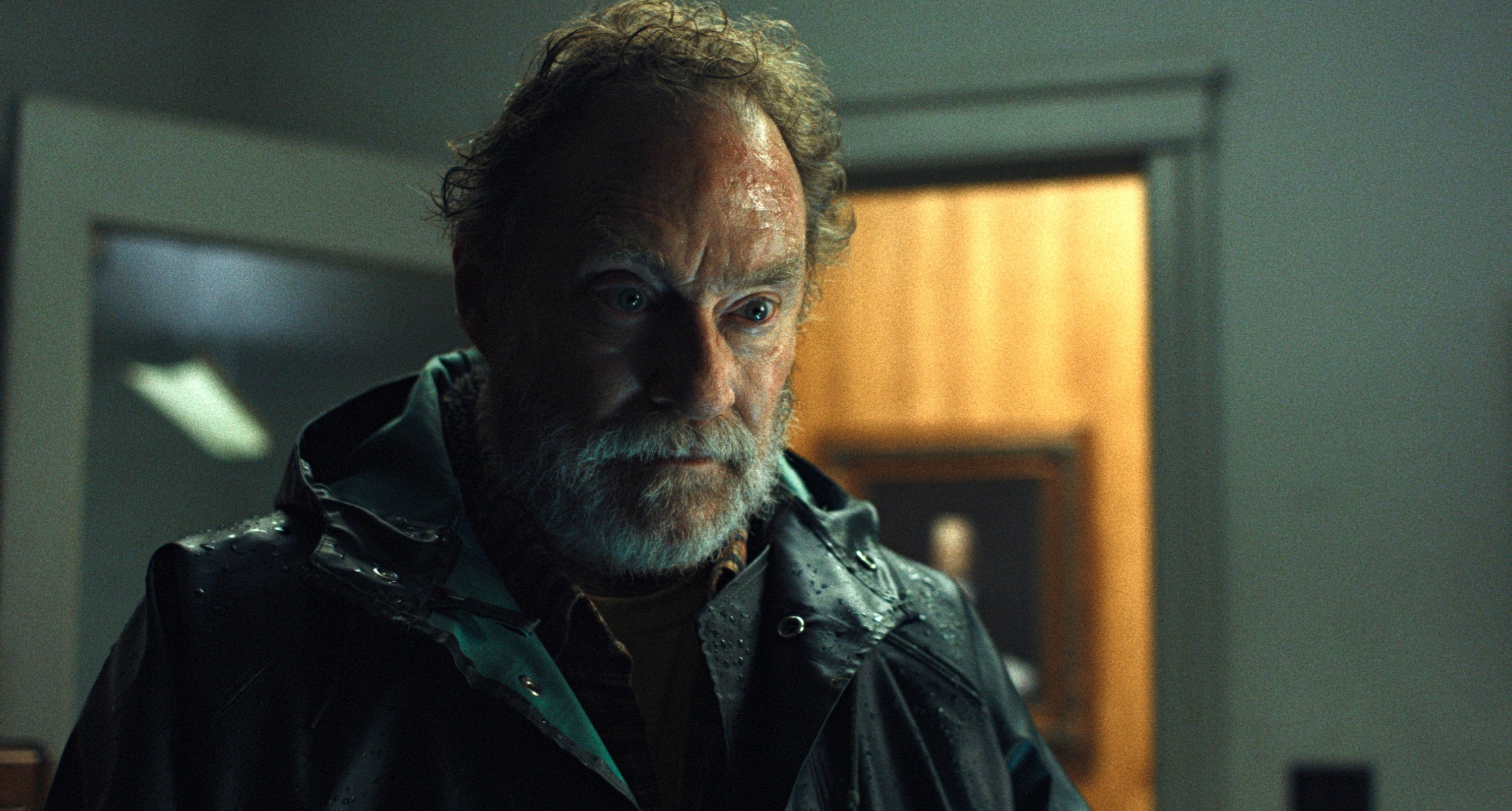 A serious-looking antheral   with a beard stands indoors, wearing a acheronian  raincoat, appearing focused and tense