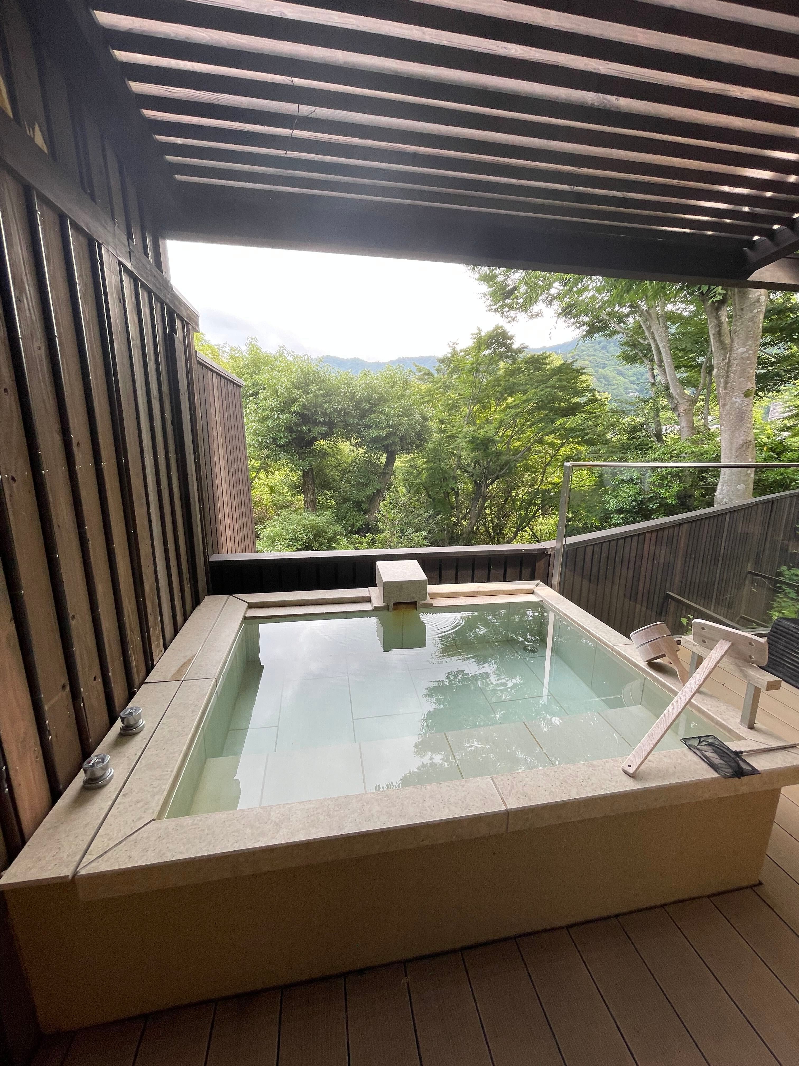 屋外にある木製の囲い付きの温泉風呂。背景に緑の木々が見える。リラックスできる、静かな雰囲気のある場所。