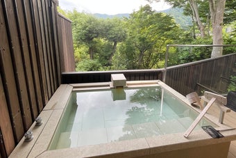 屋外にある木製の囲い付きの温泉風呂。背景に緑の木々が見える。リラックスできる、静かな雰囲気のある場所。